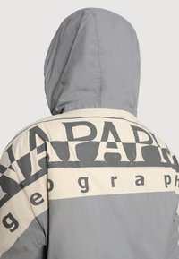 Grå huva jacka med en krämfärgad panel och stora svarta bokstäver. Tyget ser slätt ut med en subtil glans.