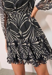 Rochie neagră și crem cu model cu frunze, material plisat, talie elastică și tiv cu volane. Designul este completat de mâneci lungi din material transparent.