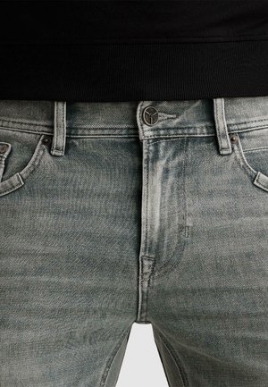 Close-up van lichtgrijze spijkerbroek met knoop- en ritssluiting, riemlussen en voorzakken, gedragen met een zwart shirt.