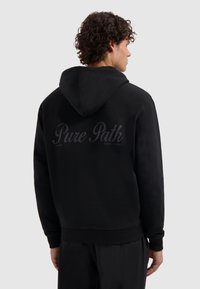 Man met krullend haar in een zwarte hoodie met de capuchon naar beneden, waarbij de achterkant de tekst "Pure Path" in donkere letters laat zien op een effen lichte achtergrond.