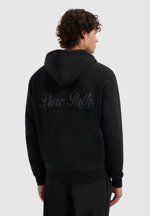 Man met krullend haar in een zwarte hoodie met de capuchon naar beneden, waarbij de achterkant de tekst "Pure Path" in donkere letters laat zien op een effen lichte achtergrond.