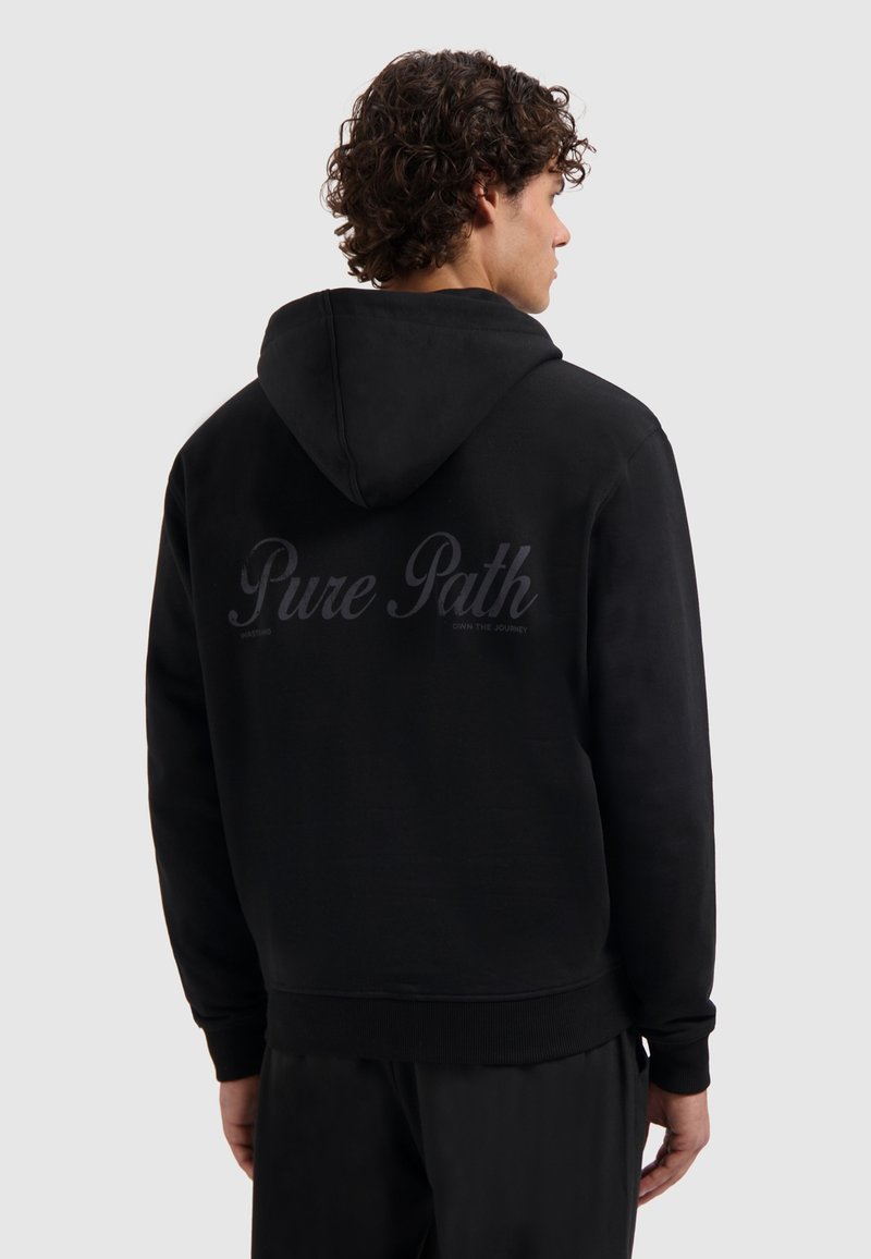 Man met krullend haar in een zwarte hoodie met de capuchon naar beneden, waarbij de achterkant de tekst "Pure Path" in donkere letters laat zien op een effen lichte achtergrond.