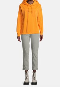 Oranžová mikina s kapucí s nápisem vpředu, šedé zúžené kalhoty a béžové šněrovací boty s tlustou černou podrážkou. Hladké textury po celém oblečení.