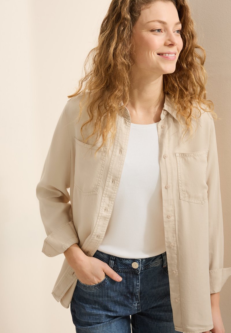 Camisa beige de botones sobre una blusa blanca, combinada con jeans de rayas verticales azul oscuro, que cuentan con dos bolsillos en el pecho y mangas remangadas.
