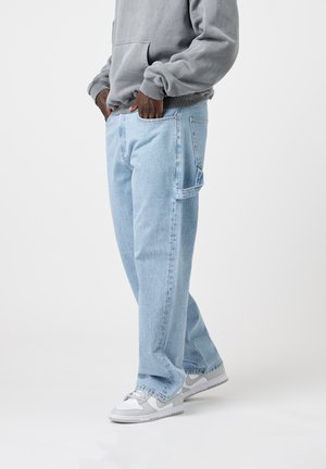 Personne portant un jean large bleu clair, un sweat gris et des baskets blanches, debout les mains dans les poches devant un fond uni.