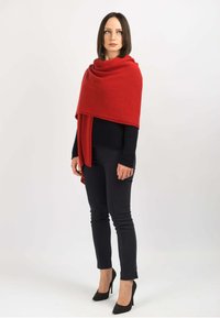 Rød uldne sjal draperet over en mørk sweater. Modellen bærer sorte skinny jeans og spidse hæle med nitter.