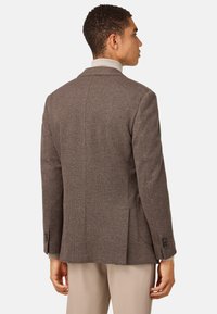 Brun texturerad kavaj med kammade slag, enkel sprund bak och två knappar vid ärmarna. Bärs över en ljusgrå tröja.