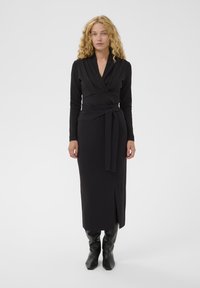 Sieviete ar lokainiem blondiem matiem, ģērbusies garā melnā apģērba ar jostas saišu un melnām augstiem zābakiem, stāvot pret baltu fona.