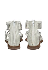 White textured flat sandals with back zippers, ankle straps featuring rhinestone embellishments, and brown soles, shown from the rear.