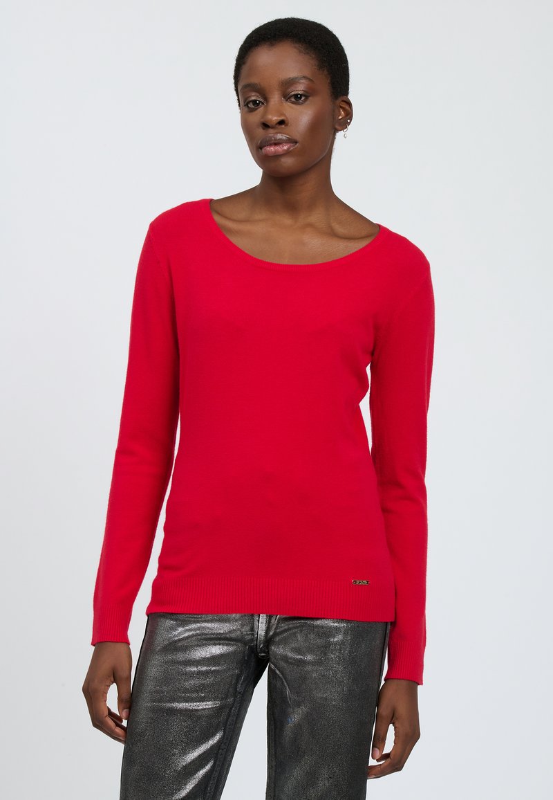 Maglione rosso a maniche lunghe con scollo a barca e orlo a coste, abbinato a pantaloni metallici argento lucidi. Texture a maglia liscia e design aderente.
