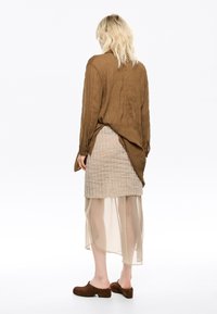 Camisa de manga larga texturizada marrón que cae sobre una falda acampanada beige con una superposición translúcida y zuecos marrones. La parte trasera muestra detalles de tela en capas.