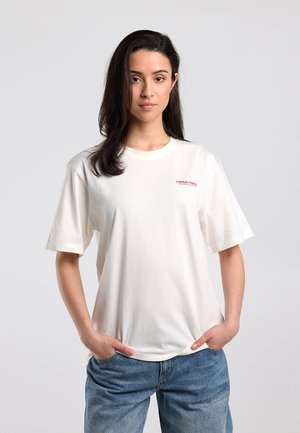 Young woman with long dark hair wearing a white oversized T-shirt with small red text and blue jeans, standing with hands in pockets.