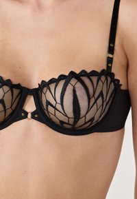 Czarny biustonosz z przezroczystymi miseczkami w kwiatowy wzór, falbankami i metalowymi detalami. Zawiera regulowane ramiączka.