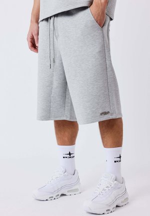 Man draagt lichtgrijze knielange shorts, witte sportsokken met zwart logo en witte sportschoenen, staat met één hand in zijn zak.