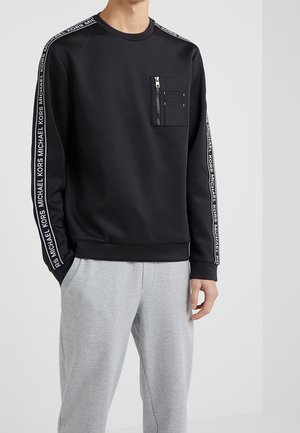Mann trägt schwarzen Sweatshirt mit langen Ärmeln mit „Michael Kors“-Logo-Band an den Ärmeln und Reißverschlusstasche auf der Brust, kombiniert mit hellgrauen Jogginghosen.