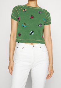 Suéter corto verde de punto con mangas cortas, decorado con insectos bordados en colores y acentos dorados a rayas en el escote y el dobladillo.