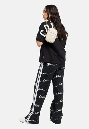 Vrouw met lang donker haar die zwarte broek met print draagt, een zwart oversized overhemd, witte sneakers en een kleine beige crossbodytas met patroon.