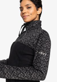 Pull-over en polaire noir et gris avec col montant, présentant un motif géométrique sur les manches et un accent logo sur le bras.