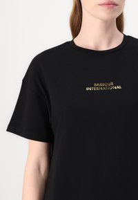 Fekete pamut póló rövid ujjakkal, kerek nyakkal, elöl arany "Barbour International" logóval.