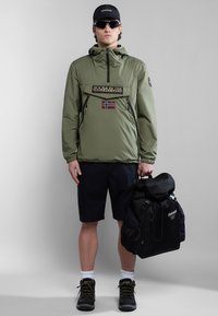 Joven con chaqueta con capucha verde oliva, pantalones cortos negros, gafas de sol y gorra, sosteniendo una mochila negra frente a un fondo gris liso.