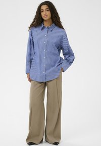 Blå og hvid stribet langærmet skjorte med krave og knaplukning fortil, kombineret med brune vide bukser.