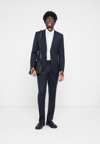 Costum bleumarin, croit ajustat, purtat cu o cămașă albă. O pereche de pantofi din piele negri și o geantă messenger neagră completează look-ul.