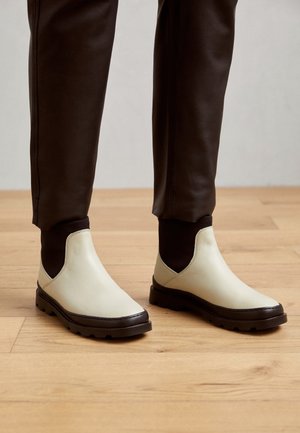 Bottines en cuir crème et marron foncé portées avec un pantalon marron foncé sur un sol en bois clair.