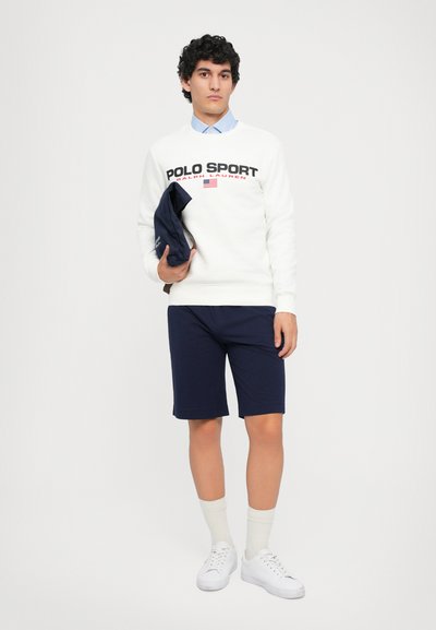 Bărbat îmbrăcat cu un hanorac alb Polo Sport, pantaloni scurți bleumarin, șosete albe și adidași, ținând o jachetă bleumarin pliată, pe un fundal neutru.