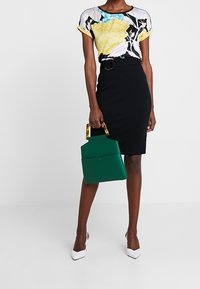 Květovaný krátký top s krátkými rukávy v černé, žluté a tyrkysové; černá pouzdrová sukně; zelená kabelka s geometrickým tvarem; bílé boty na vysokém podpatku.