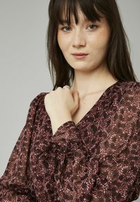 Blouse maroon à motifs floraux avec de longues manches ballon, tissu texturé et poignets froncés, présentant un design à col en V profond.