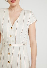 White dress with vertical brown stripes, V-neck, short sleeves, and gold buttons. Features a belt at the waist and pleats at the skirt.