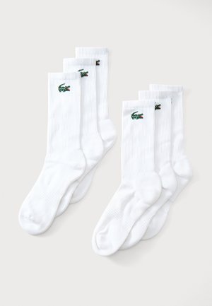Zwei Sets weiße Crew-Socken mit grün gesticktem Krokodil-Logo, nebeneinander auf hellem Hintergrund präsentiert.