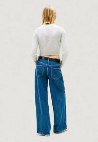 Maglione grigio corto e jeans denim blu a gamba larga con grandi tasche posteriori. Presenta una cintura nera con borchie argentate.