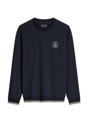 Jeune homme portant un sweat-shirt bleu marine avec bordure blanche et logo, short bleu marine avec le numéro 67 imprimé, chaussettes blanches et baskets blanches.