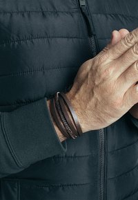 Pulseras de cuero marrón en una muñeca, con diferentes texturas y grosores, apiladas sobre una mano contra un fondo de chaqueta con cremallera oscura.