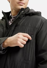 Czarna izolowana kurtka z zamkniętą kieszonką na zamek na klatce piersiowej. Posiada kaptur i gładką fakturę. Model dostosowuje zamek w kieszeni.