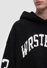 Musta neulehuppari, jossa on ribbikuviointi, huppu ja rohkeaa valkoista tekstiä, jossa lukee "WRSTB". Mukana valkoiset yksityiskohdat hihoissa.