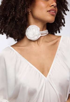 Collana choker bianca con un design floreale testurizzato e accenti sottili, abbinata a una blusa bianca con scollatura a V e texture plissettata.