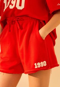 Røde bomuldsshorts med elastisk talje, der har snor, sidelommer og et hvidt "1990" trykt på kanten.