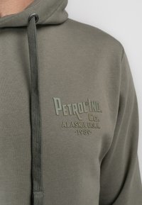 Primer plano de una sudadera verde oliva con "Petrol Ind. Co. Alaska U.S.A. 1989" impreso en el pecho, con un cordón con ojales de metal.