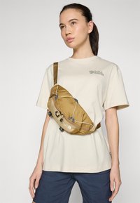 Žlto-hnedá crossbody taška s nastaviteľným popruhom, textúrovanou tkaninou, elastickými šnúrkami a kovovými akcentmi, nosená cez svetlo béžovú košeľu.