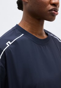 Κοντινό πλάνο ενός ατόμου που φοράει μπλε σκούρο πουκάμισο με λευκές γεωμετρικές γραμμές στους ώμους, με φόντο απλό και ανοιχτόχρωμο.