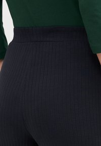 Strakke, ribbelige donkerblauwe broek gedragen met een donkergroene lange mouw top, waarbij details rond de taille en heupen zichtbaar zijn.