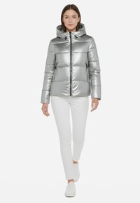 Giacca a vento argentata con una texture lucida e riflettente, chiusura centrale con zip, cappuccio e tasche laterali, abbinata a pantaloni bianchi e sneakers.