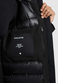 Käsi hoiab avatud musta polsterdatud jakki, mille sees on silt, kus on kirjas "KHUJOTM Genuine Brand Established 2001" ja väike vahtramärgi ikoon.