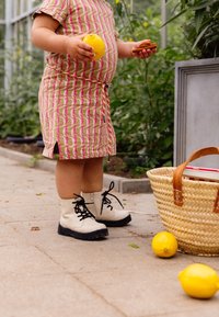 Una niña vestida con un vestido de estampado y botas sostiene un limón y una galleta, con una cesta de mimbre y dos limones en el suelo cerca.