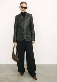 Blazer in pelle nera con chiusura a bottoni, abbinato a pantaloni neri a gamba larga e scarpe beige a punta con texture. Accessoriato con una borsa marrone e occhiali da sole.