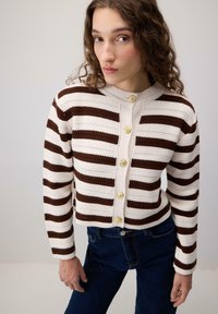 Femeie îmbrăcată într-un cardigan cu dungi orizontale albe și maro, cu butoane aurii, și blugi de culoare albastru închis, stând în fața unui fundal simplu.