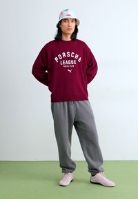 Burgunderrød sweatshirt med hvid "PORSCHE LEAGUE" tekst, grå sweatpants og lyserøde sneakers. Modellen bærer en pastelbucket hat. Enkel baggrund.