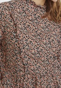 Blommig blus med volangkrage, rosa och vita små blommor på en mörk bakgrund samt en rynkad midja.